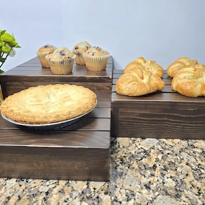 May include: Two dark brown wooden tiered display risers holding baked goods. The lower tier holds a round pie in a metal tin, while the upper tier holds four muffins and two golden-brown croissants. A glass vase with a red rose and yellow flowers.
