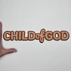 May include: A beige patch with the words "CHILD of GOD" in a stylized font. The letters have a red outline and a textured, fuzzy appearance. The patch is likely for clothing or accessories.