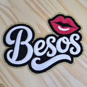May include: A patch with the word "Besos" in white script lettering on a black background, outlined with gold glitter. A red lip graphic is in the upper right corner.