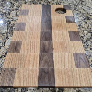 May include: A rectangular wooden cutting board with a mosaic pattern of light and dark brown wood pieces. A circular hole is cut into one corner. The board is made of various wood grains and has a natural finish.