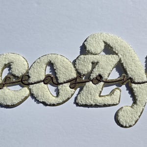 May include: A decorative sign spelling "cozy" in a fluffy, off-white material with a gold border. The word "season" is written in a cursive font across the word "cozy". The sign is on a white background.