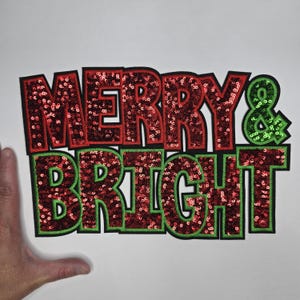 May include: A festive patch with the words "MERRY & BRIGHT" in red sequined letters outlined in black and green. The ampersand is green. Perfect for holiday crafts and apparel.