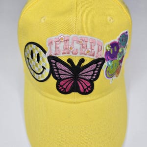 May include: Bright yellow baseball cap with embroidered patches. Patches include a smiley face, a butterfly, and the word "TEACHER" in pink letters. Additional patches feature colorful smiley faces. The cap is made of a durable fabric.