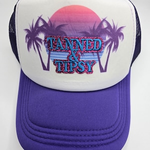 May include: A purple and white trucker hat with the words "TANNED & TIPSY" in glittery blue and pink lettering. The hat features a sunset graphic with palm trees. The brim is purple, and the mesh backing is navy blue.