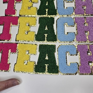 Peut inclure: Gros plan sur des lettres en feutre colorées épelant "TEACH". Les lettres sont roses, jaunes, vertes, bleu clair et violettes, avec des bordures dorées à paillettes. Les lettres sont disposées sur une surface blanche.