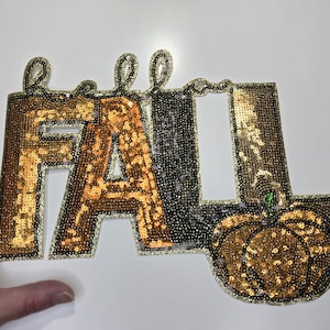 May include: A sequined "Hello Fall" sign with gold, black, and silver sequins. The word "Fall" is in gold sequins, and the pumpkin is also gold. The sign is a fall-themed decoration.
