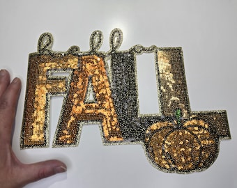 May include: A sequined "Hello Fall" sign with gold, black, and silver sequins. The word "Fall" is in gold sequins, and the pumpkin is also gold. The sign is a fall-themed decoration.