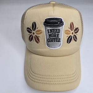 May include: A tan trucker hat with a mesh back and a curved peak. The front displays a coffee cup graphic with the text "I NEED MORE COFFEE" and floral designs. The hat is a neutral colour, suitable for everyday wear.