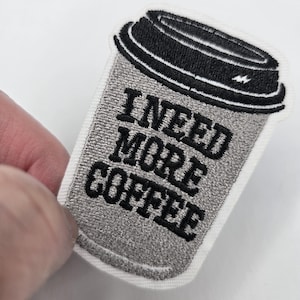 May include: Embroidered patch in the shape of a coffee cup, with a black lid and gray cup. The text "I NEED MORE COFFEE" is embroidered in black on the cup. The patch is held by a hand.