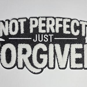 May include: A black and white decorative sign with the words "NOT PERFECT JUST FORGIVEN" in a bold, textured font. The text is layered on a black background with a white border, creating a striking contrast. The sign is likely for home decor.