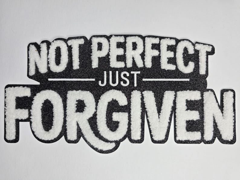 May include: A black and white decorative sign with the words "NOT PERFECT JUST FORGIVEN" in a bold, textured font. The text is layered on a black background with a white border, creating a striking contrast. The sign is likely for home decor.