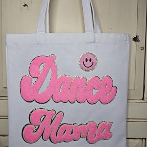 May include: White canvas tote bag with the words "Dance Mama" in pink, outlined in gold glitter. A small, round, gold glitter patch with a smiling face is also on the bag. The tote bag has white handles.