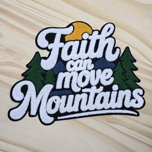 May include: A white, black, and green patch with the text "Faith can move Mountains". The patch features a yellow sun, green trees, and blue mountains. The patch is on a light brown wooden surface.