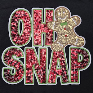 May include: A black graphic with the words "OH SNAP" in red sequins with a green outline and white border. A gold sequined gingerbread man with a green bow tie is on the right side.