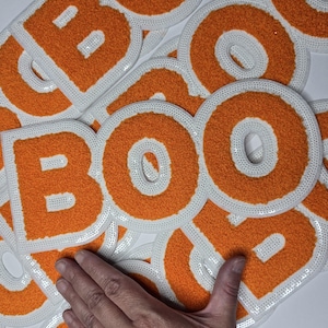 May include: Orange felt letter patches spelling "BOO" with white sequin borders. The patches are scattered across a white surface. The letters are in a varsity style.