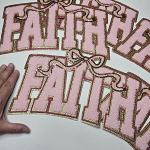 May include: Pink fabric patches spelling "FAITH" in a collegiate font. The letters have gold trim and are topped with sequined bow accents. The patches are on a white surface.