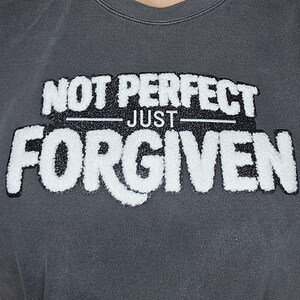 May include: Dark gray sweatshirt with the text "NOT PERFECT JUST FORGIVEN" in a white, textured font. The sweatshirt has a crew neck and a relaxed fit, suitable for casual wear.