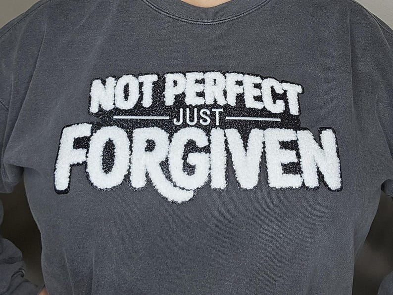 May include: Dark gray sweatshirt with the text "NOT PERFECT JUST FORGIVEN" in a white, textured font. The sweatshirt has a crew neck and a relaxed fit, suitable for casual wear.