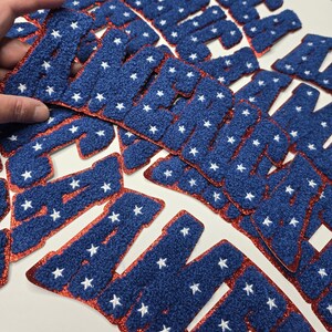 May include: Blue, star-studded felt letters with a red glitter border. The letters are arranged in a pile, with one being held by a hand. The letters appear to spell out a word, possibly a name or a phrase.