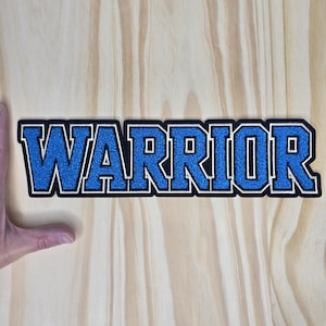 May include: A blue and white embroidered patch with the word "WARRIOR" in bold, block letters. The patch has a black border and is set against a light wood grain background.