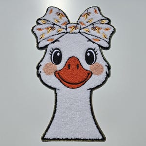 May include: A white goose patch with a large bow featuring a pattern of gold and orange leaves. The goose has a bright orange beak, rosy cheeks, and large black eyes. The patch has a black border.