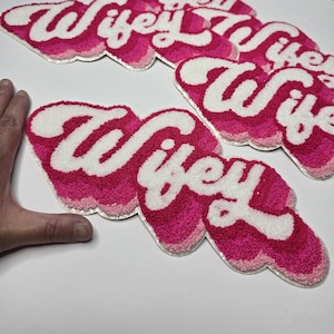 May include: Several pink and white "Wifey" patches with a cloud-like border. The patches have a layered design with a white top layer, a medium pink middle layer, and a darker pink bottom layer. The word "Wifey" is written in a cursive font.