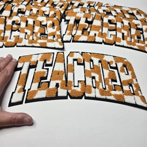 May include: Several "TEACHER" patches with a white and orange chequered design. The letters are thick and textured, with a felt-like appearance and a black border. The patches are displayed on a white surface.