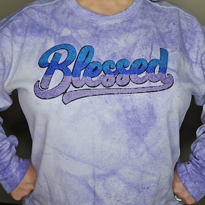 May include: A light purple tie-dye sweatshirt with the word "Blessed" in blue and purple glitter lettering. The sweatshirt has long sleeves and a crew neck. The overall style is casual and comfortable.