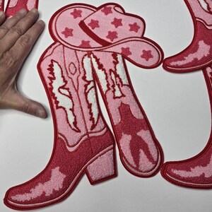 May include: Pink and red cowboy boot and hat patches. The boots have a detailed design with white accents and a block heel. The hat is decorated with red star shapes. The patches are made of a textured material.