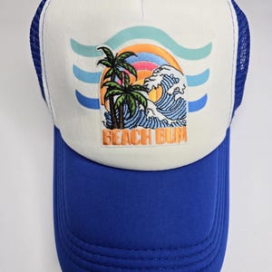 Peut inclure: Casquette de camionneur bleue et blanche avec un écusson brodé sur le thème de la plage. L'écusson représente des palmiers, un coucher de soleil et des vagues océaniques, avec le texte "BEACH BUM". La casquette a une visière bleue et un dos en filet.