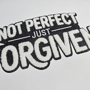 May include: A black and white patch with the words "NOT PERFECT" above "JUST FORGIVEN" in a bold, textured font. The patch has a glittery black background and a white border.