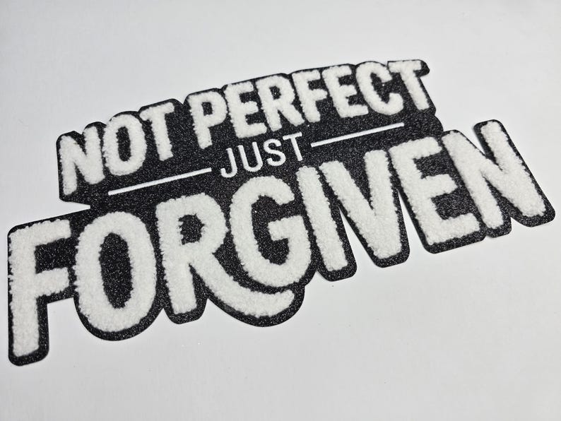 May include: A black and white patch with the words "NOT PERFECT" above "JUST FORGIVEN" in a bold, textured font. The patch has a glittery black background and a white border.