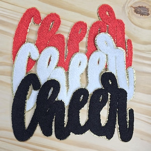 May include: Close-up of the word "Cheer" in red, white, and black, with gold trim. The letters are made of a textured material, possibly felt or fabric, and are arranged in a layered design. The background is a light wood surface.