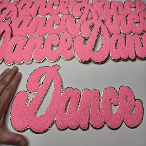 May include: Pink, fuzzy "Dance" word cutouts with gold glitter edging. The word is in a cursive font. Several cutouts are visible, suggesting a craft or party decoration theme.