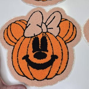 May include: Orange and black pumpkin-shaped rug with a smiling face and mouse ears, resembling a popular cartoon character. The rug has a light pink border and a textured appearance, perfect for Halloween decor.