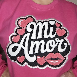 May include: Pink sweatshirt with the phrase "Mi Amor" in white, black, and red lettering, surrounded by hearts and a pair of lips. The design has a silver glitter outline.