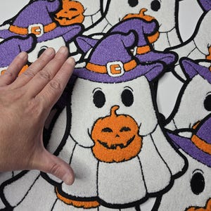 May include: White ghost-shaped Halloween decorations with black outlines. Each ghost wears a purple witch hat with an orange band and a smiling orange pumpkin. The decorations are made of a soft, textured material.