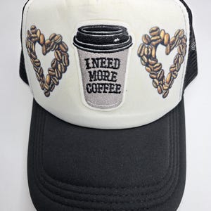 May include: Black and white trucker hat with a coffee theme. The hat features a white front panel with a coffee cup graphic and the text "I NEED MORE COFFEE." Two heart shapes made of coffee beans flank the cup.