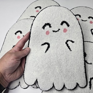 May include: White ghost-shaped rugs with black outlines and smiling faces with pink cheeks. The rugs have a textured surface and a scalloped bottom edge. The image shows several of these rugs, with one being held by a hand.