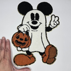 May include: A Halloween-themed patch featuring a cartoon character dressed as a ghost, holding a jack-o'-lantern. The character has black ears, a white body, and orange shoes. The patch is outlined in gold.