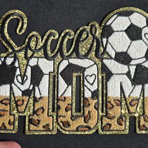 May include: A black and gold glitter patch with the words "Soccer MOM". The word "Soccer" is in gold glitter script. The letters in "MOM" are filled with a leopard print design. The soccer ball and hearts are white and black.