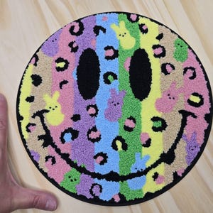 May include: A round, colorful rug with a smiley face design. The rug features a rainbow of colors, including pink, purple, blue, green, and yellow, with black leopard print spots and bunny shapes. The eyes and mouth are black.
