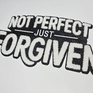 May include: A black and white patch with the words "NOT PERFECT JUST FORGIVEN". The text is in a bold, stylized font with a fuzzy, textured appearance. The patch is on a white background.
