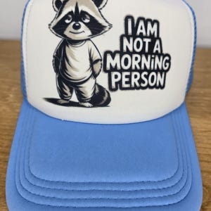 May include: A light blue and white trucker hat with a cartoon raccoon graphic. The raccoon is wearing a white shirt and has its hands in its pockets. The text "I AM NOT A MORNING PERSON" is printed on the front.