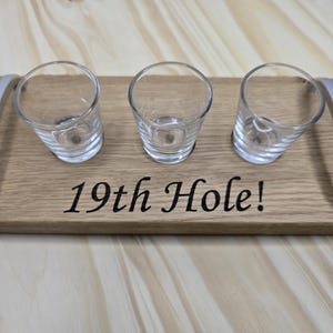 May include: A wooden serving tray with three shot glasses. The tray has silver handles and the words "19th Hole!" are printed on the wood. The shot glasses are clear and cylindrical.