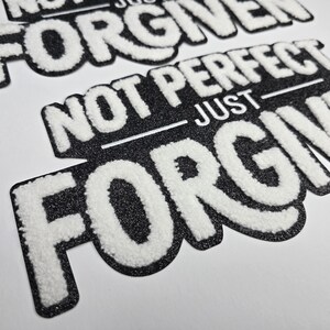 May include: Close-up of black and white patches with the text "NOT PERFECT JUST FORGIVEN". The text is in a bold, textured font with a white, fluffy appearance, set against a black, glittery background.
