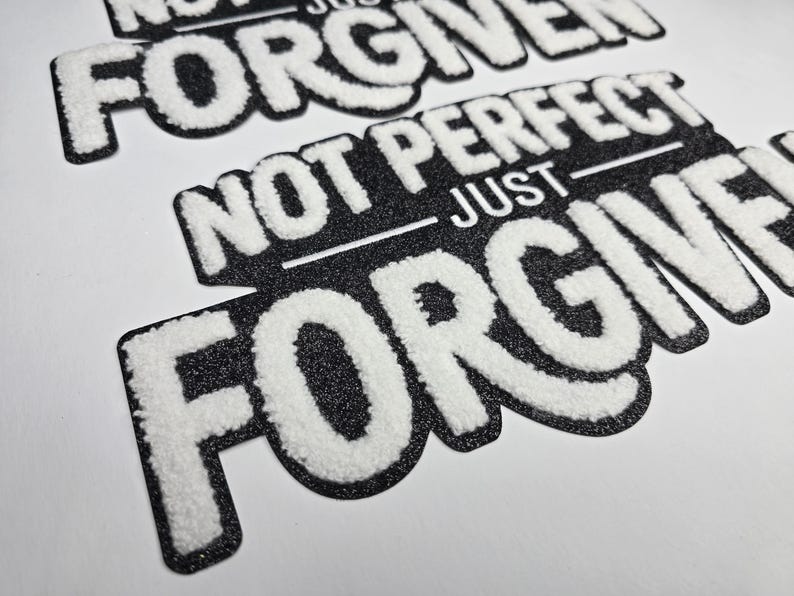 May include: Close-up of black and white patches with the text "NOT PERFECT JUST FORGIVEN". The text is in a bold, textured font with a white, fluffy appearance, set against a black, glittery background.