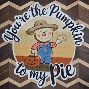 May include: A round, colorful rug with a fall theme. The rug features a smiling scarecrow holding a slice of pie and a jack-o'-lantern. Text on the rug reads "You're the Pumpkin to my Pie". The background has a chevron pattern.