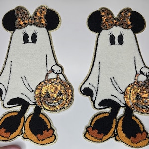 May include: Two Halloween-themed patches featuring a ghost-like Minnie Mouse design. The patches have black ears, a sequined bow, and a pumpkin-shaped treat bag. The design is outlined in gold and has black and orange accents.