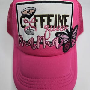 May include: A bright pink trucker hat with a mesh back and a curved brim. The front features a white rectangle with a cocktail illustration and the words "Caffeine Queen Mama" in embroidered lettering, along with a butterfly applique.